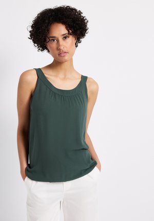 Dark green sleeveless top with a rounded neckline, subtle pleating at the front, and a smooth, flowing fabric. Paired with white pants.
