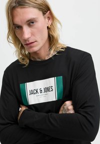 Blonder Mann mit Tattoos und goldenem Ohrring trägt ein schwarzes Langarmshirt mit dem Logo "JACK & JONES COLLECTIONS EST. 1990" und hat die Arme verschränkt.