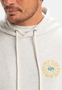 Lys grå hættetrøje med en frontlomme, hvide snøresnore og et farverigt logo-design på brystet med gule og grønne detaljer.