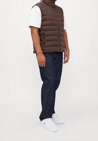 Brun puffer väst med hög krage, dragkedja framtill och sidofickor, i kombination med en vit t-shirt och mörka denimjeans, avslutat med vita sneakers.