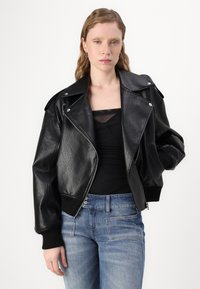 Giacca bomber in pelle nera con finitura testurizzata, ampi risvolti e bottoni a pressione argentati, abbinata a una maglietta nera e jeans blu.