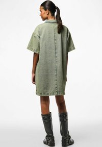 Preveliki zeleni traper haljina sa kratkim rukavima, ovratnikom i bočnim džepovima, uparen s crnim čizmama do koljena na kopčanje.