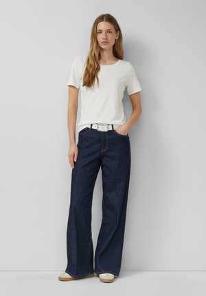 Vrouw met lang haar, die een wit t-shirt, witte riem, donkere wijd uitlopende spijkerbroek en witte sneakers draagt, staand tegen een simpele grijze muur.