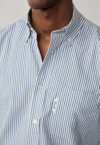 Camisa de botón a rayas en algodón de color azul claro y blanco, con cuello, un bolsillo en el pecho con una etiqueta y botones blancos. Tejido de textura fina.