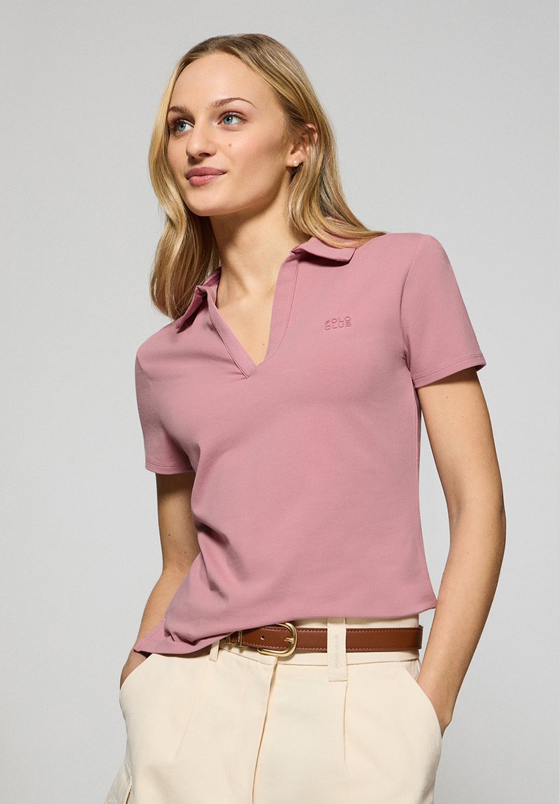 Rosa polo skjorta med v-ringning och korta ärmar, tillverkad av mjukt tyg. Logotyp på vänster bröst. I kombination med beige byxor.