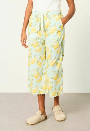 Frau trägt hellblaue Hosen mit gelbem Zitronenprint, wellenförmigen Säumen und einem elastischen Bund, kombiniert mit beigen Slippern.