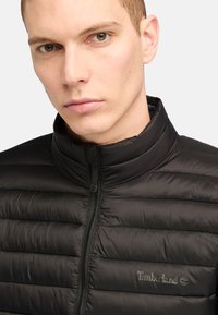 Giubbotto imbottito nero con collo alto, dotato di motivi trapuntati, chiusura frontale con zip e logo Timberland in rilievo sul petto.