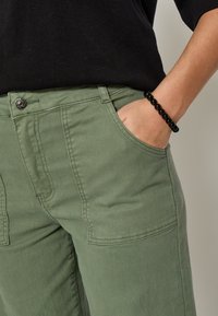 Cilvēks, kuram ir olīvu zaļas bikses ar priekšējiem kabatām un melns pērlīšu aproce, roka atpūšas labajā kabatā.