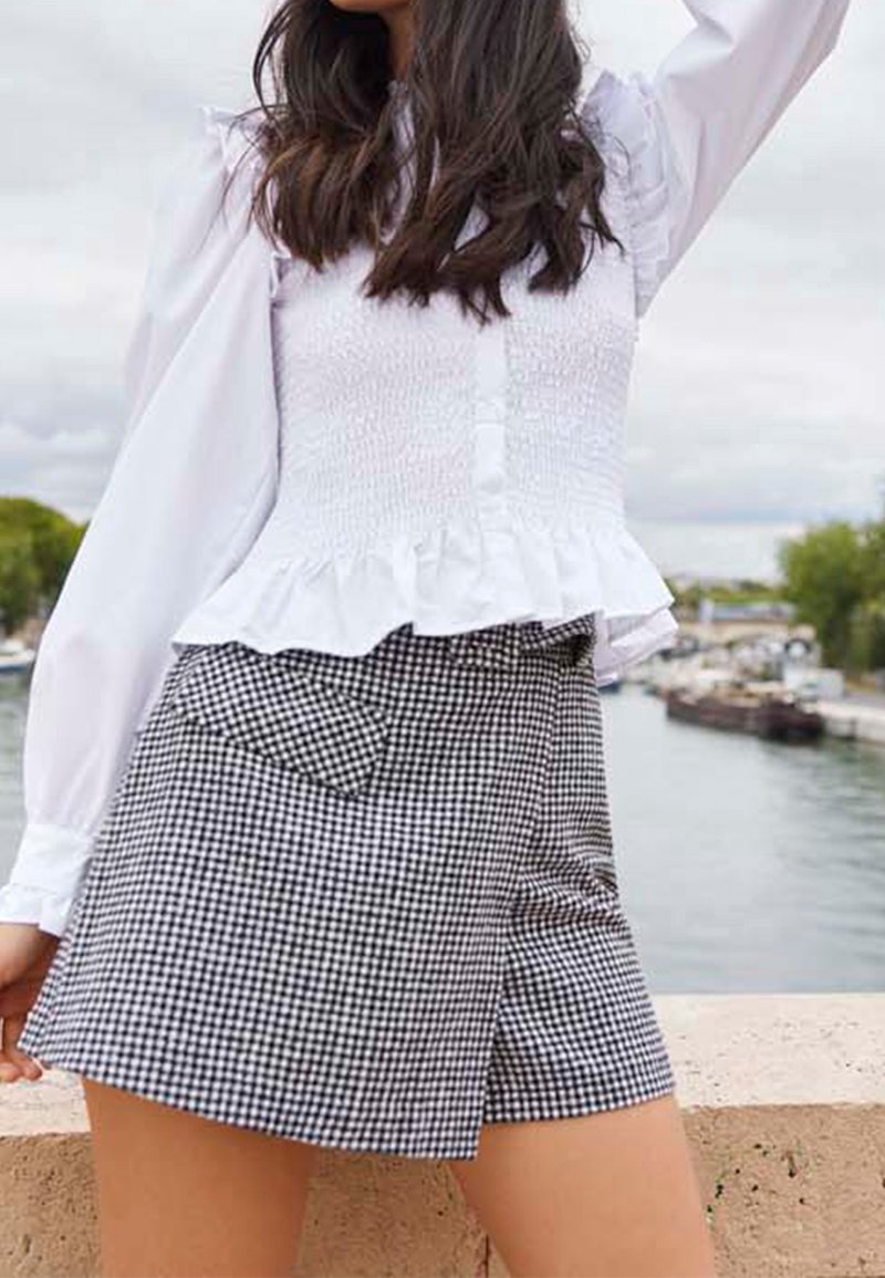 Kvinna klädd i vit volangblus och svart-vitrutig kjol som står vid en flod med båtar och träd i bakgrunden.