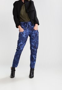 Blå byxor i kamouflagemönster med avslappnad passform, snörning i midjan och sidfickor, ihop med en svart jacka och ankelstövlar.