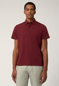 Burgundská polo košeľa s krátkymi rukávmi, golierom a malým vyšitým logom na hrudi, spárovaná s svetlými ľanovými nohavicami.