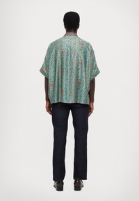 Kortærmet skjorte i turkis med intrikate mønsterdetaljer. Stoffet ser let og luftigt ud og har en afslappet pasform, parret med mørke jeans.
