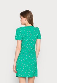 Vestido floral verde com mangas curtas bufantes, apresentando padrões de flores brancas e vermelhas, uma silhueta ajustada e fecho de zíper nas costas.