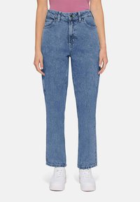 Vista frontal de uma pessoa a usar jeans retos de cintura alta azuis e ténis brancos sobre um fundo simples.