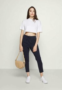 Vit kortärmad t-shirt, marinblå joggers med dragsko och vita sneakers. Håller en beige texturerad axelväska. Enkel, modern design.