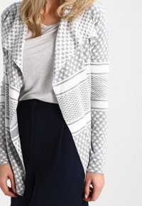 Šedý a bílý vzorovaný cardigan s otevřeným předním dílem, obsahující geometrické vzory a dlouhé rukávy, spárovaný se světle šedým topem a tmavými kalhotami.