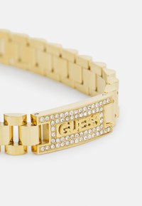 Bracelet en métal doré au design massif, doté d'un fermoir central orné de strass transparents et du logo "GUESS". Maillons texturés.