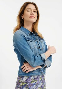 Svijetloplavi traper jakna s dva džepa na prsima, kopčama na dugmad i zarolanim rukavima, nosi se preko haljine s cvjetnim uzorkom.