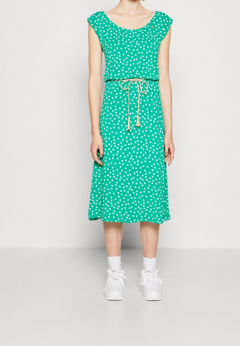 Robe verte à pois avec un col rond, des manches courtes et une taille cintrée nouée avec un cordon beige orné de pompons.