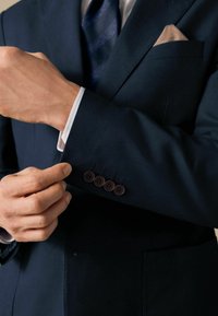 Marineblauer Blazer mit glatter Textur, ausgestattet mit vier braunen Knöpfen an den Manschetten und einem cremefarbenen Taschentuch im Brusttasche.