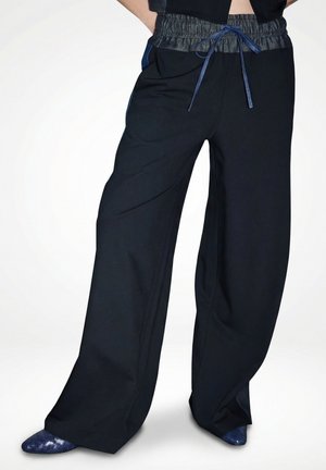 Weit geschnittene schwarze Hose mit elastischem Bund und blauem Kordelzug, kombiniert mit spitzen dunkelblauen gewebten Schuhen.