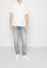 Camisa blanca de manga corta con diseños bordados recortados, combinada con jeans de color azul claro y zapatillas blancas.