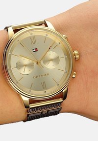 Montre-bracelet dorée avec un cadran rond, face texturée, chiffres romains, petits compteurs et un bracelet en métal maillé. Comprend le logo de la marque.