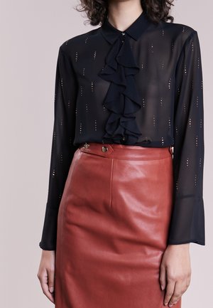 Femme portant un chemisier noir transparent avec des volants et des pois métalliques, associé à une jupe en cuir marron taille haute avec des détails de boutons.