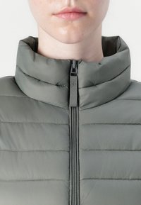 Giacca imbottita grigia con colletto alto e zip frontale. Il tessuto ha una texture liscia con un motivo di cuciture orizzontali per l'isolamento.