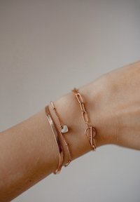 Tre braccialetti in oro rosa su un polso: uno con un ciondolo a forma di cuore, un bracciale piatto texture e un bracciale a catena con un dettaglio a chiusura a gancio.