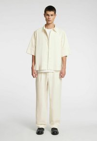 Kremowa koszula na krótki rękaw z zapięciem na guziki z przodu, w połączeniu z pasującymi luźnymi spodniami i czarnymi butami.