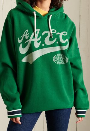 Sudadera verde con un gran logotipo blanco "A.E.C." y un logotipo más pequeño en la parte inferior. Tiene cordones y puños a rayas. Tejido suave.