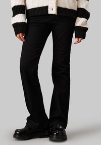 Schwarze, weit geschnittene Jeans aus einem glatten Material, mit einem klassischen Fünf-Taschen-Design, kombiniert mit klobigen schwarzen Schuhen und einer gerippten Strickjacke.