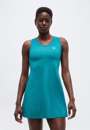 Urheilullinen nainen, joka pukeutunut hihattomaan turkoosiin urheilumekkoon ja seisoo yksivärisen vaalean taustan edessä.