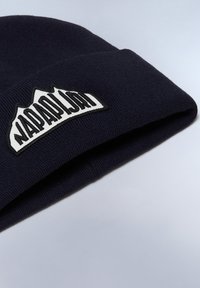 Berretto a maglia navy con risvolto. Presenta una patch bianca con lettering nero e una grafica di montagna. Texture morbida, vestibilità aderente.