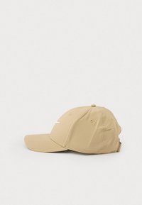 Gorra de béisbol beige con visera curva y correa ajustable, que presenta un pequeño logo en forma de swoosh blanco en el panel frontal, mostrada de lado.