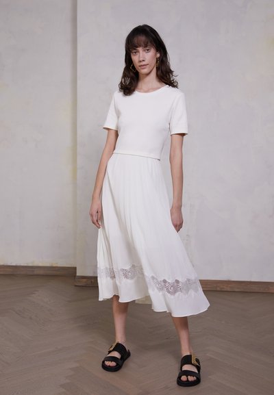 White dress with a fitted top and flared, pleated skirt. Features lace detailing at the hem. Worn with black sandals. Smooth texture.