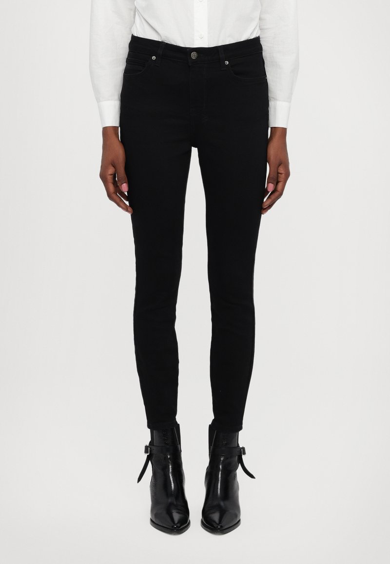 Jeans skinny negros de denim, con un diseño clásico de cinco bolsillos, cierre de botón metálico y detalles de costura. Llevados con botas negras hasta el tobillo.