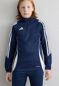 Top desportivo azul marinho com riscas brancas nas laterais e logo da Adidas, com colarinho de meia zip e mangas longas. Usado com calças a condizer.