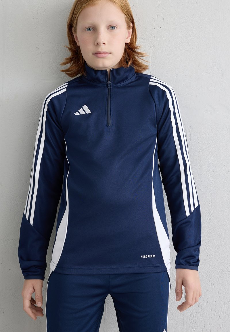 Top desportivo azul marinho com riscas brancas nas laterais e logo da Adidas, com colarinho de meia zip e mangas longas. Usado com calças a condizer.