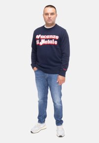 Felpa navy a collo rotondo con "Vacanze di Natale" in lettere rosse, abbinata a jeans azzurri e sneakers grigi. Design semplice, senza motivi.