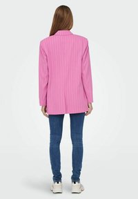 Blazer rosa a righe con una vestibilità su misura, maniche lunghe e silhouette dritta, abbinato a jeans blu e sneakers bianche.