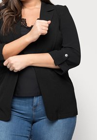 Zwarte blazer met een gestructureerd ontwerp, textuurstof, driekwartmouwen en zilveren knoopaccenten. Gedragen over een zwart topje en blauwe jeans.