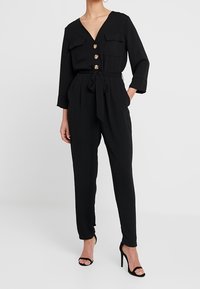 Svart jumpsuit med V-ringning, framfickor, knappar och knyt i midjan. Tyget har en mjuk struktur. Bärs med remsandaler.