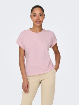 Vrouw staat met hand in de zak, draagt een roze-wit gestreept T-shirt met korte mouwen en beige broek tegen een effen achtergrond.