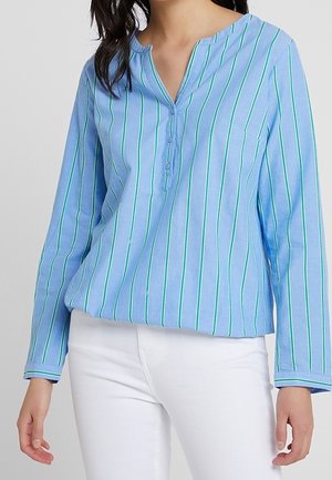 Jasnoniebieska bluzka z długim rękawem w pionowe zielone i białe paski, zapięta przy szyi, noszona z białymi spodniami.