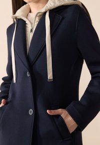 Cappotto blu navy con un design aderente, caratterizzato da un cappuccio foderato in beige. Include tasche frontali, una chiusura a bottoni e lacci di colore chiaro.