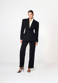 Costum negru, croi atletic, cu un sacou structurat, cu pad-uri ascuțite la umeri și pantaloni largi; suprapus peste un top crem. Tocurile completează look-ul.