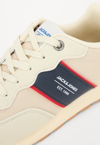 Beige joggesko med mesh og glatte materialer, marineblå og røde detaljer, runde snørehull og logo på siden.
