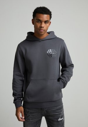 Jeune homme portant un sweat à capuche gris foncé avec une poche avant et un logo sur la poitrine, associé à un jean noir déchiré, debout devant un fond uni.